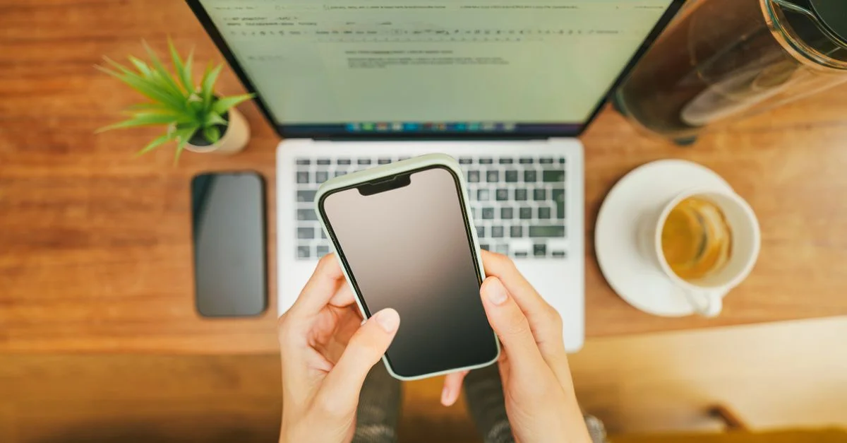 Mains tenant un smartphone sur un ordinateur portable avec du café dans un environnement de bureau à domicile confortable.